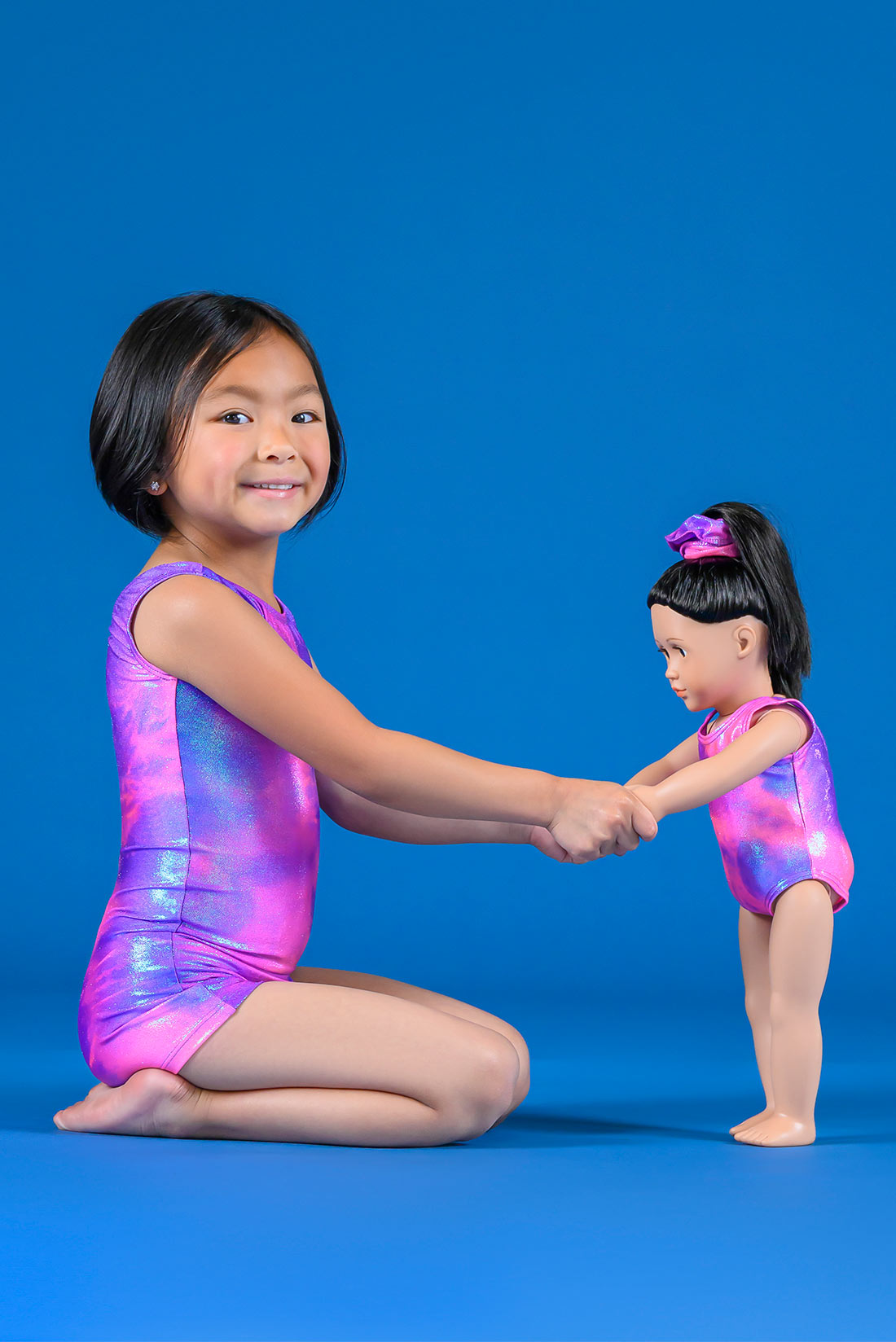 A toddler girl wearing a gymnastics unitard and holding hands with a doll wearing a matching pink and purple leotard by Destira, 2023