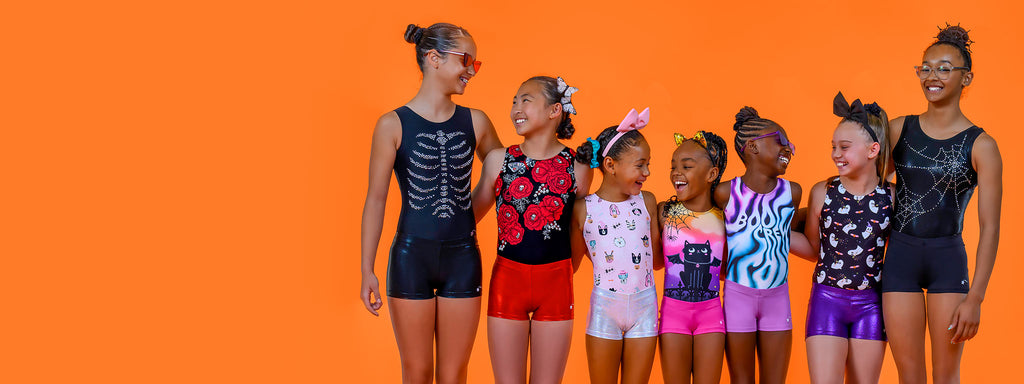 Seven girls ages 7-13 wear Halloween-themed leotards and coordinating shorts and stand with their arms around each other, laughing, in front of a solid, bright orange background.