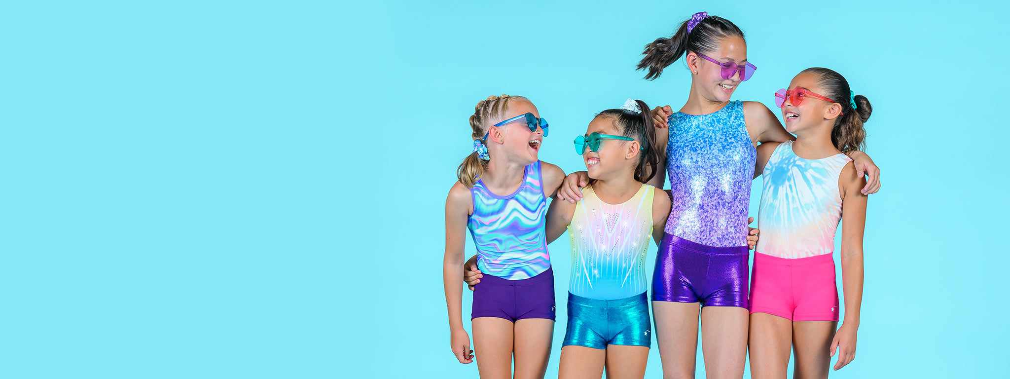 Four tween girls wearing pastel blue, purple, and rainbow leotards with pink, purple, and aqua matte and metallic shorts and jelly sunglasses stand and laugh with their arms around each other in front of a sky blue solid background.