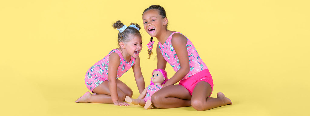 Two preschool-age girls wearing pink unicorn print gymnastics apparel sit on their knees, laughing and holding a doll wearing a matching pink leotard, in front of a solid, pale yellow background.
