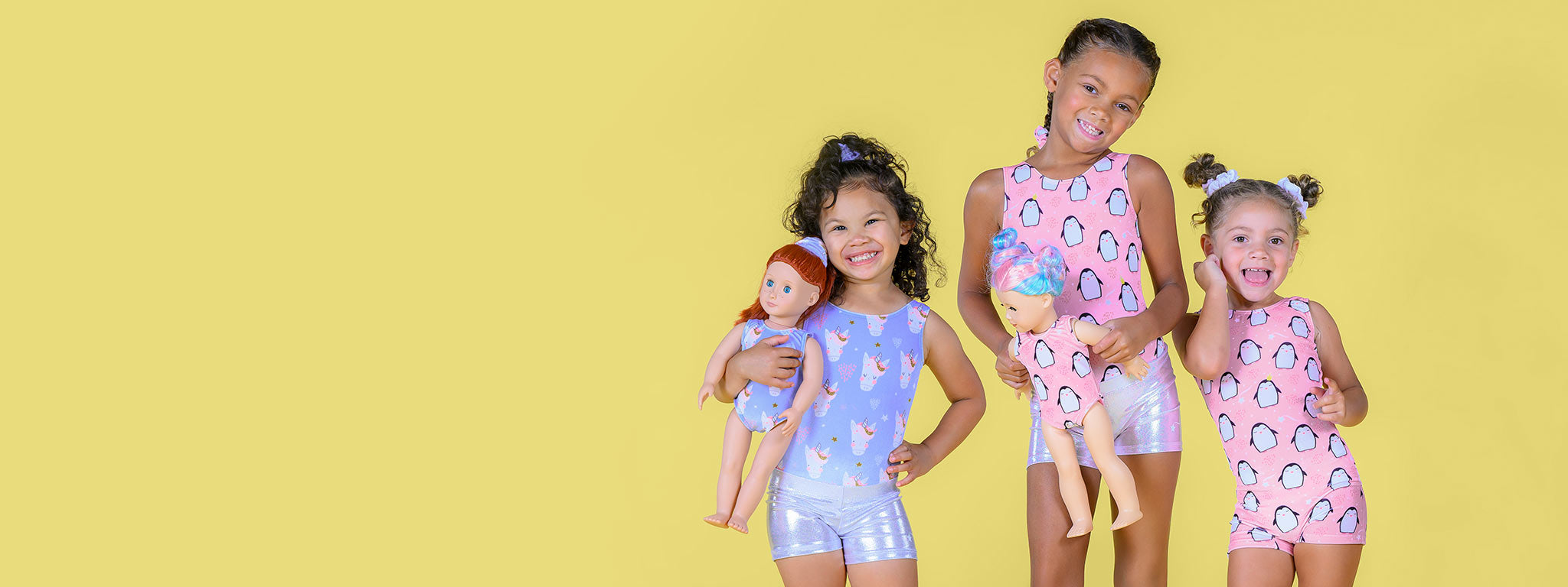 Three girls aged 5-7 years old wear velvet leotards and unitards and metallic silver shorts in front of a solid light yellow background. Two of the girls are holding dolls, which are wearing doll-sized leotards that match the girls'.