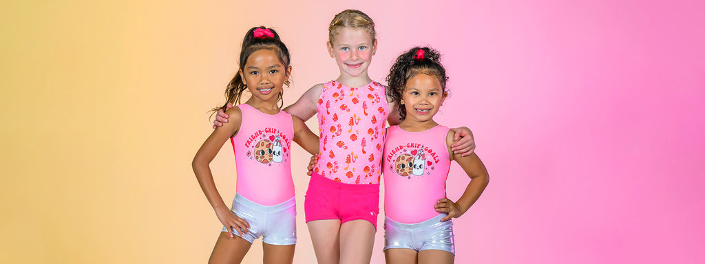 Three young girls wearing solid berry pink mystique leotards and silver mystique shorts stand smiling with their hands on their hips in front of a solid baby pink background.