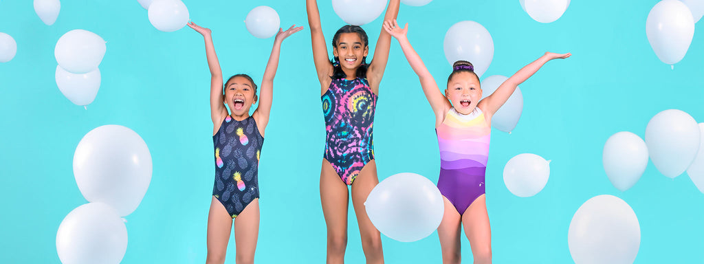 Three young girls wearing popular Destira leotards smile and throw their hands up as white balloons fall around them in front of a solid turquoise background.