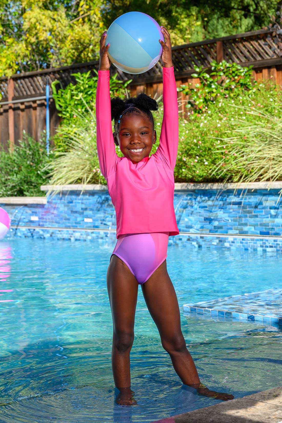 Child in pink long sleeve rash guard over colorful swimsuit holding a beach ball by a pool