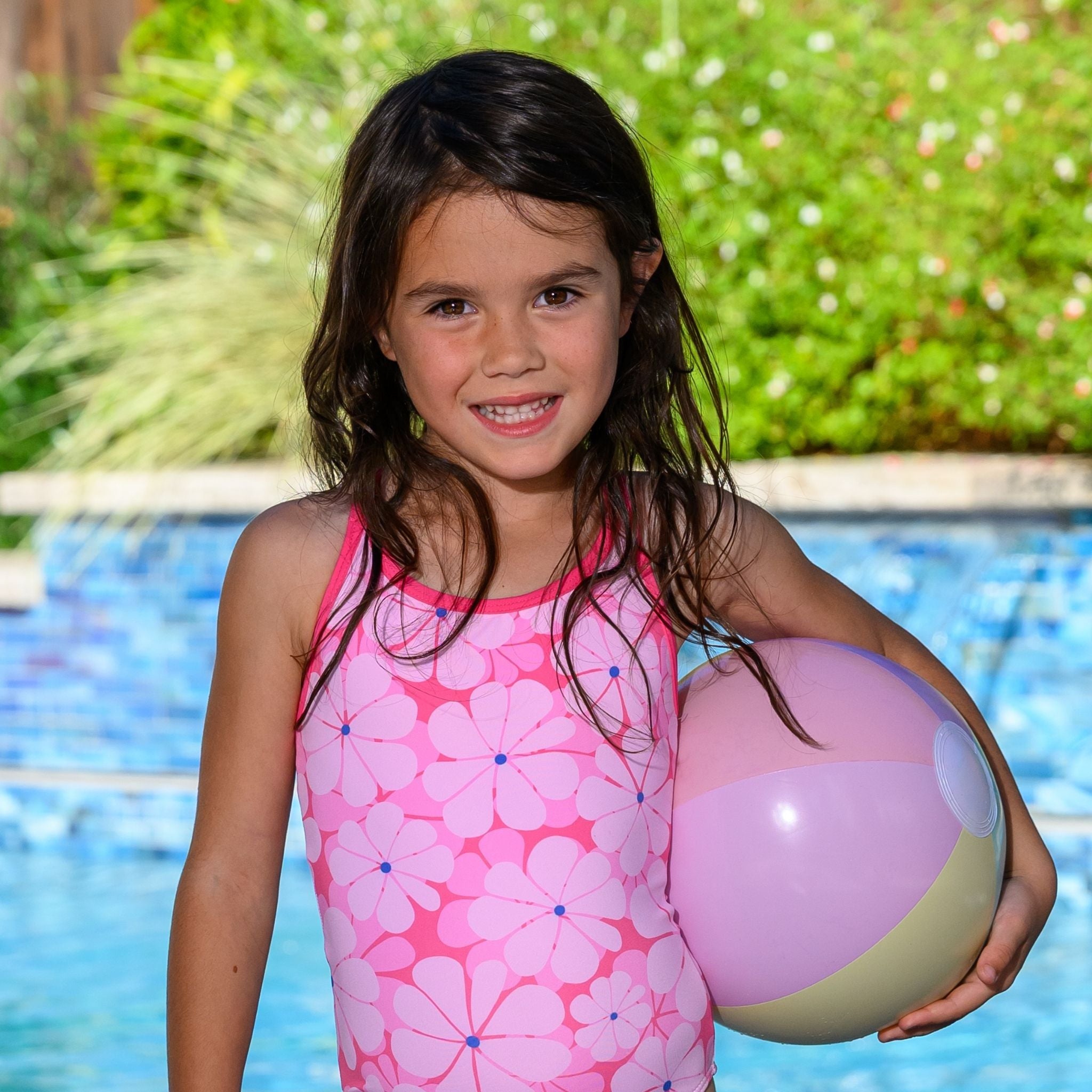 Young girl in pink floral one-piece swimsuit holding a beach ball by a pool.