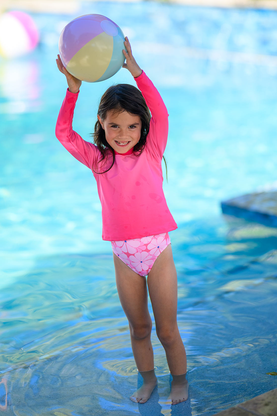 Girl in pink swimsuit with rash guard holding a colorful ball in a pool