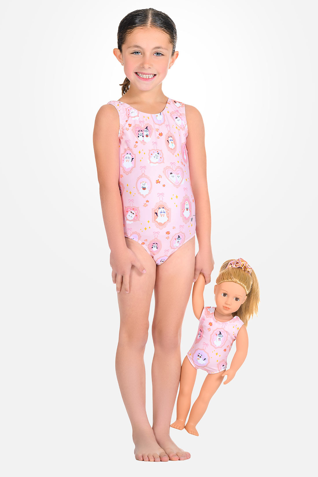 A young girl stands barefoot against a light gray studio background wearing a sleeveless pink one-piece leotard with a white and pastel cartoon print. A smaller doll in a matching leotard held beside her.
