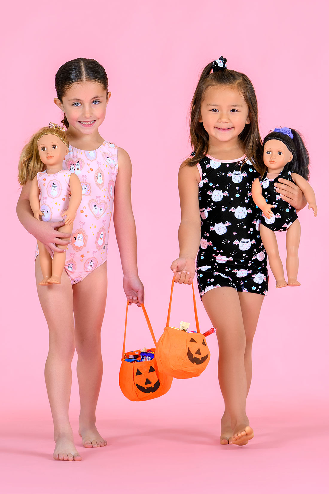 Two young girls standing against a solid pink studio background, each holding a doll and a small orange pumpkin-shaped trick-or-treat bucket. The girl on the left wears a light pink leotard with white cartoon patterns and holds a blonde doll.