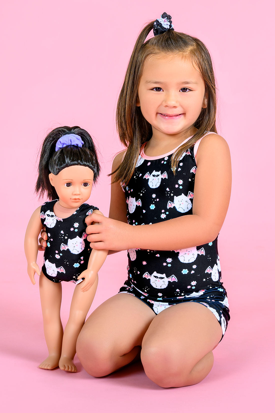A young girl kneeling on a pink background wearing a unitard with the meowloween kitten pattern, while holding a matching doll.