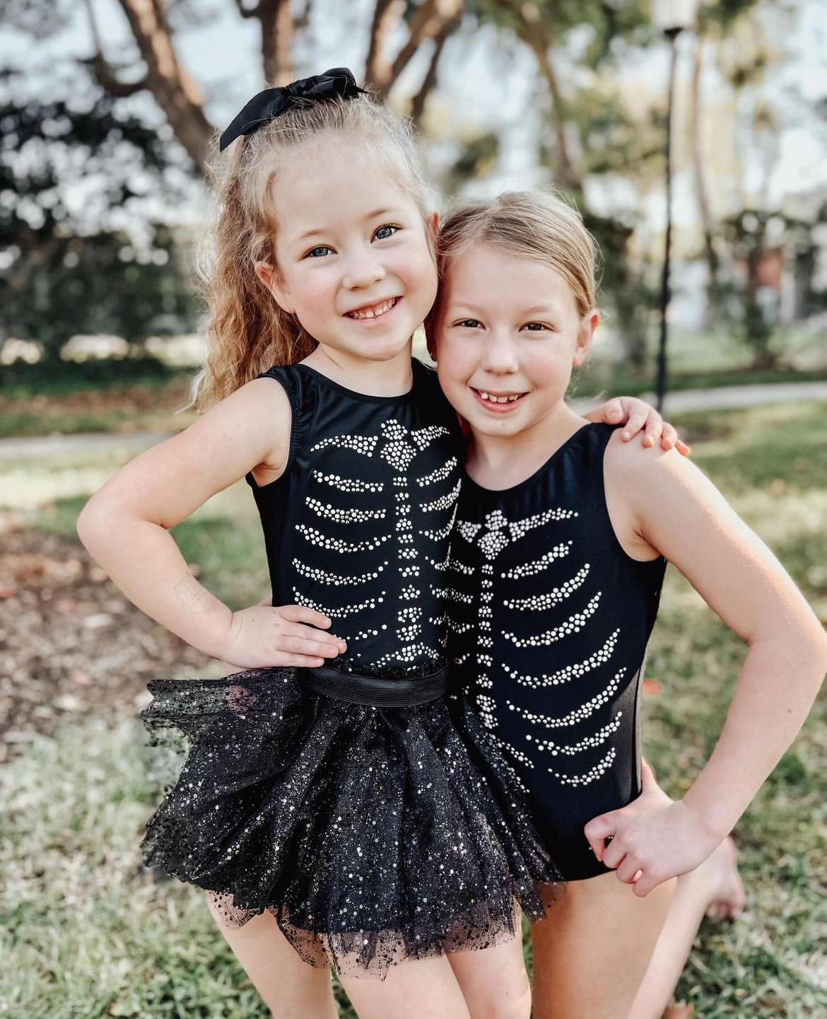 Two young girls wearing matching black sleeveless leotards with a silver rhinestone skeleton rib cage design and black tulle skirts, posing together outdoors on grass.
