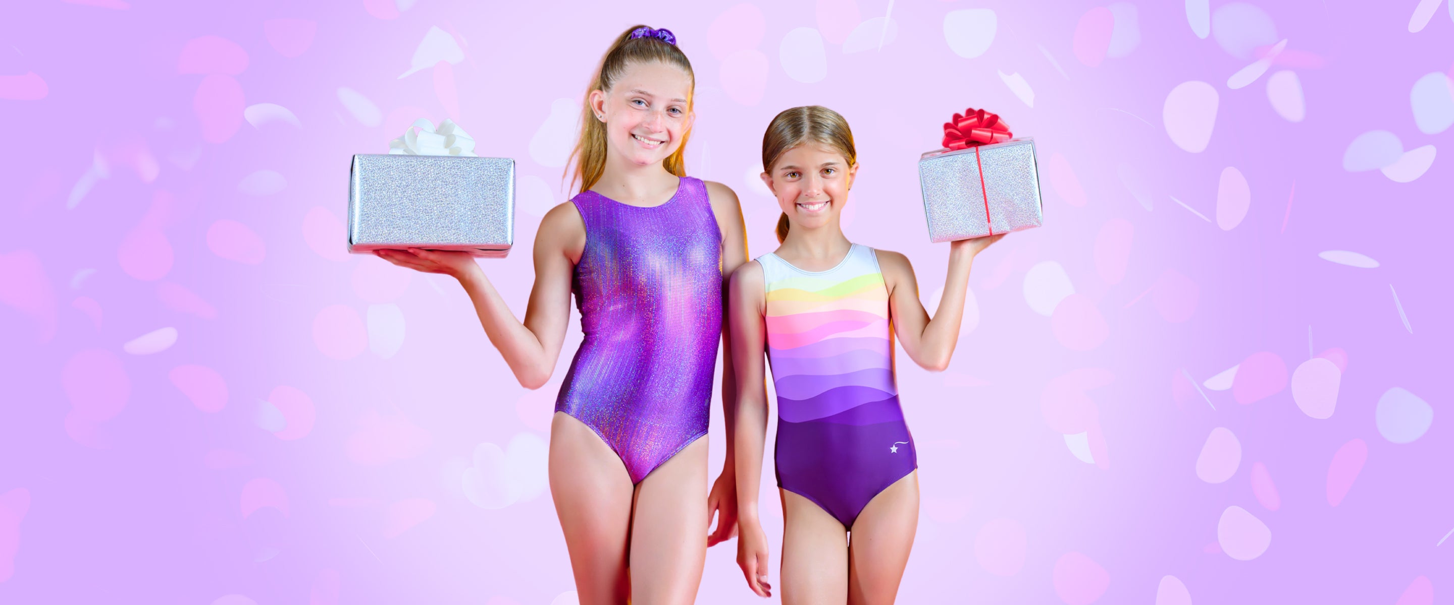 Two young girls in colorful leotards holding gift boxes against a pink background