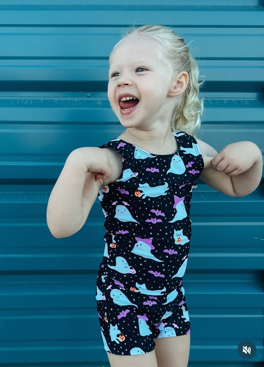 A young blonde girl wearing a black unitard with a light blue and purple ghost pattern, posing in front of a blue corrugated metal wall.