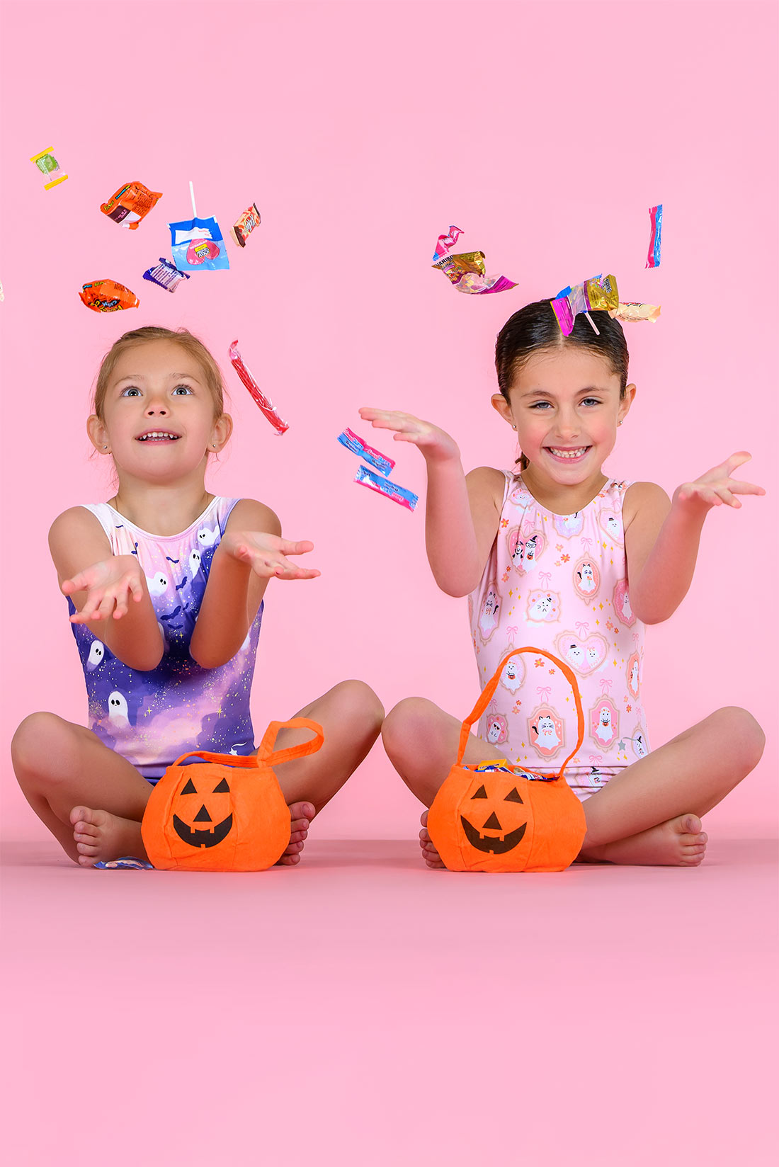 Destira Halloween gymnastics leotards. Two young girls holding orange pumpkin-shaped trick-or-treat buckets and reaching toward candy pieces floating in the air. 