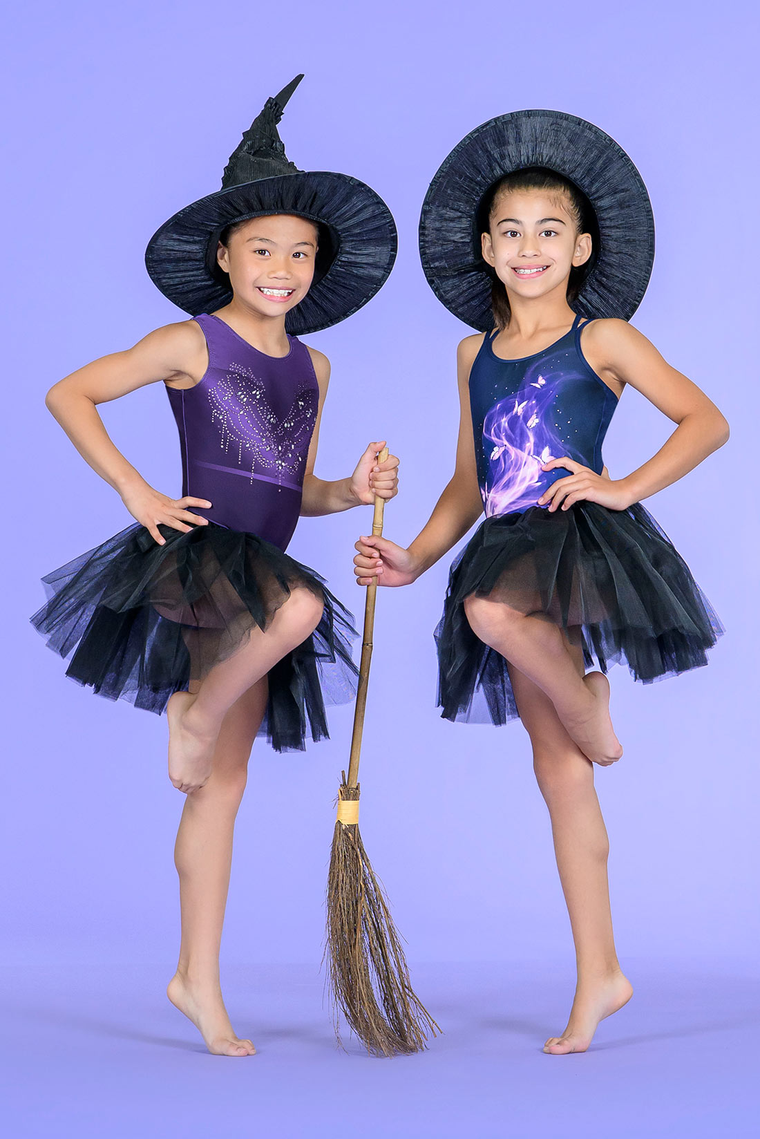 Two young girls in witch leotard costumes posing in Destira leotarda with black tutus against a solid lavender studio background. 
